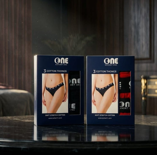 Two navy blue retail boxes of One Design One underwear packaging standing on a dark reflective tabletop in an indoor setting. The boxes show product photos and text reading '3 COTTON THONGS' and 'SOFT STRETCH COTTON.