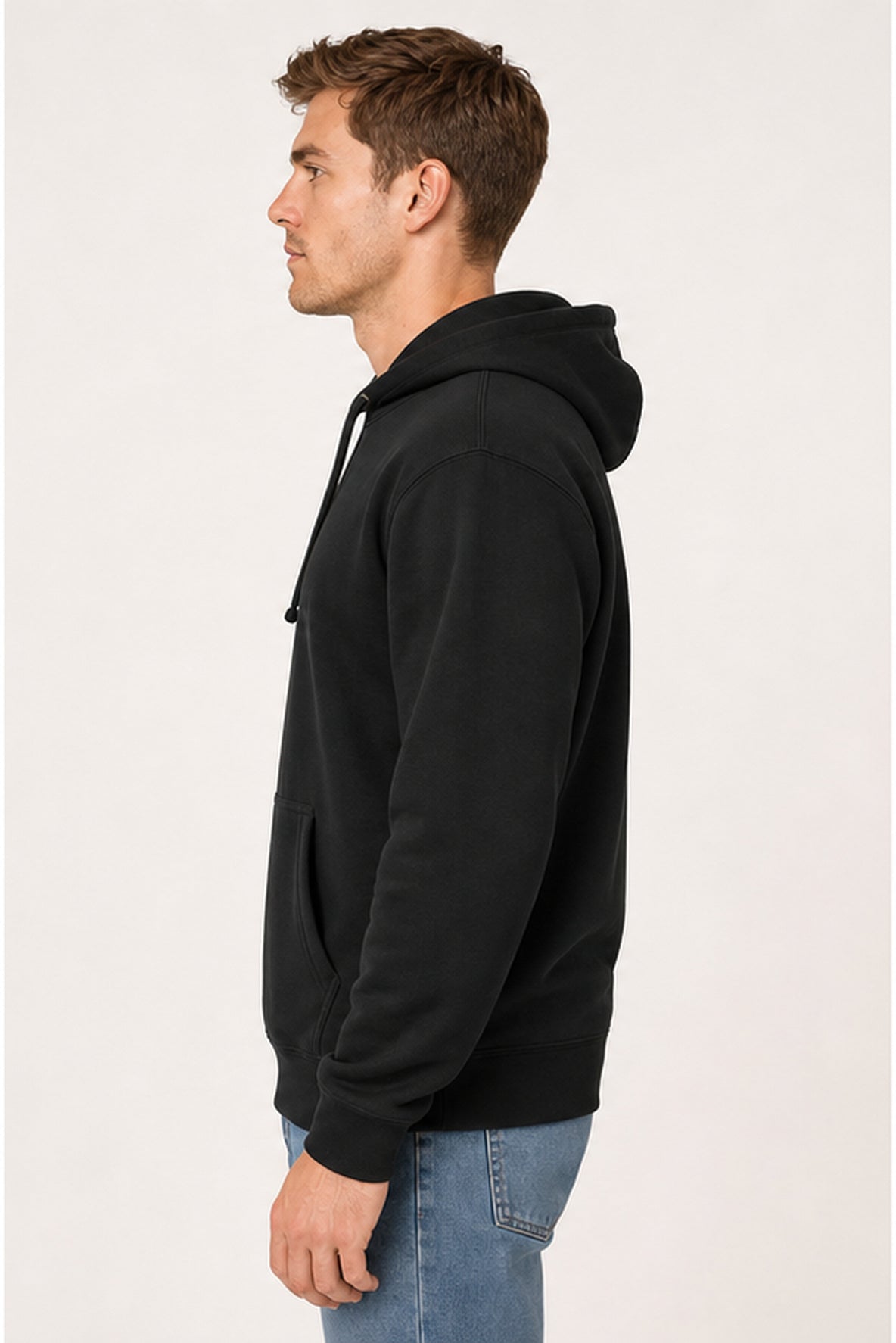 A man in profile wearing a black pullover hoodie and blue jeans, standing against a plain light background.
