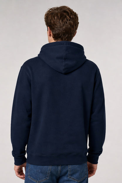 A man viewed from the back wearing a dark navy blue pullover hoodie and blue jeans against a light gray studio background.