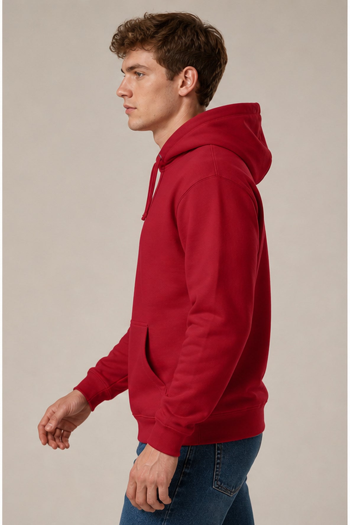 A man in profile wearing a solid red pullover hoodie and blue jeans, standing against a plain light beige studio background.