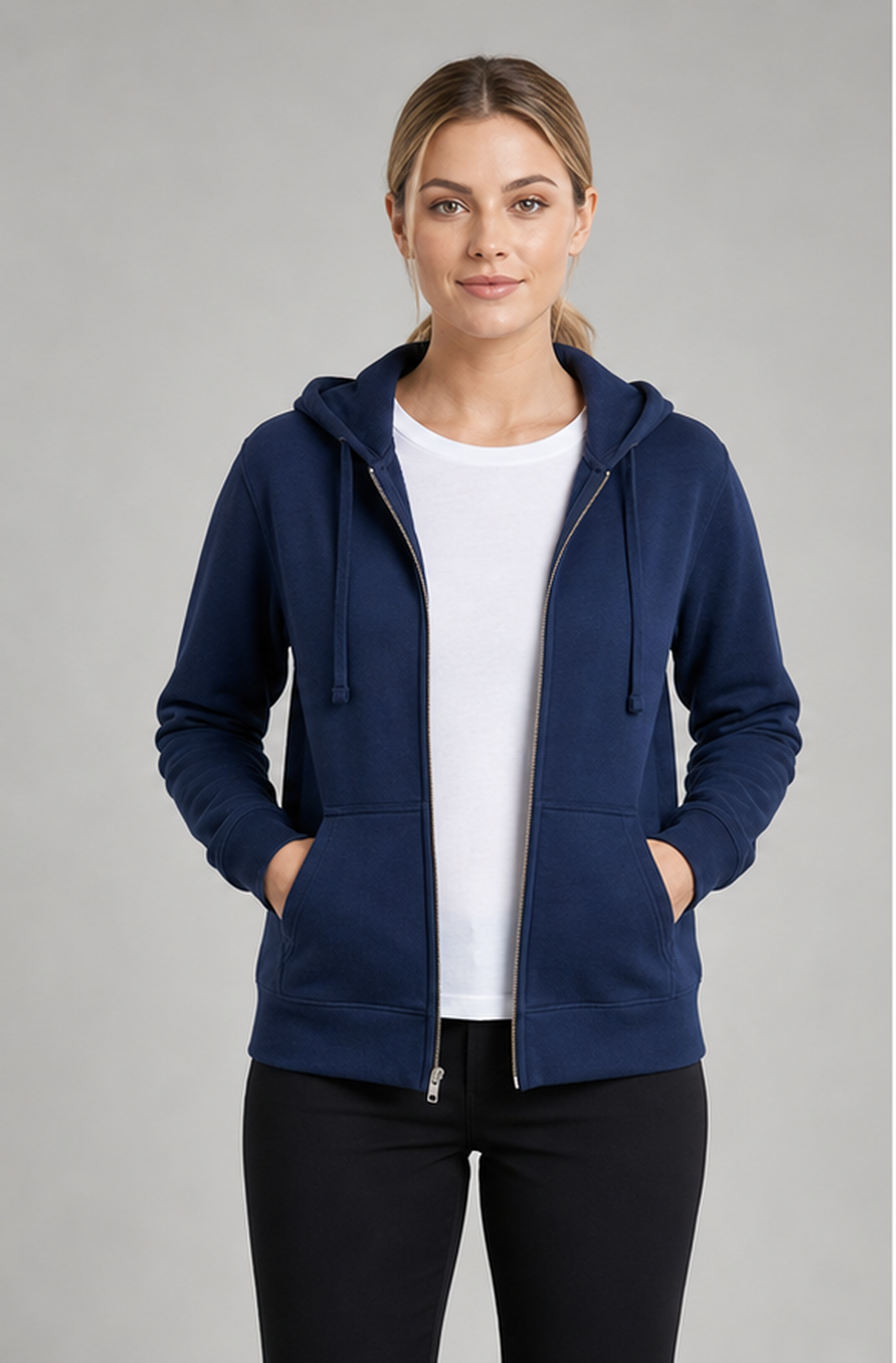 A woman modeling a navy blue zip-up hoodie with drawstrings and front pockets, worn open over a white t-shirt and black pants, against a solid gray studio background.